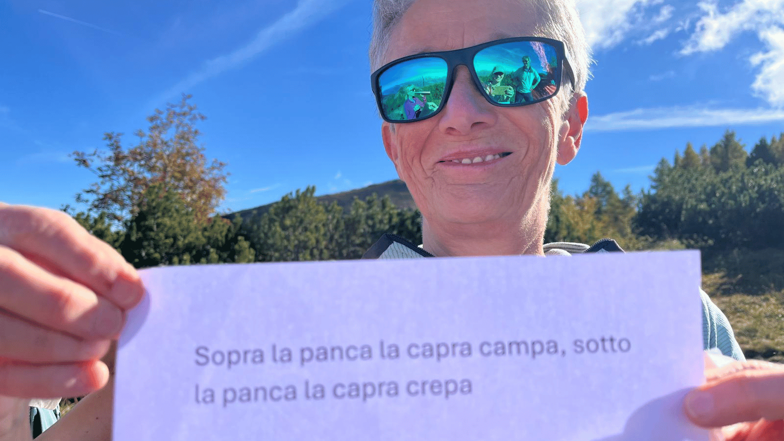 Frau mit Sonnenbrille, in der sich andere Personen spiegeln, hält ein Papier mit einem Zungenbrecher in die Kamera. Im Hintergrund Wald und ein grüner Berggipfel