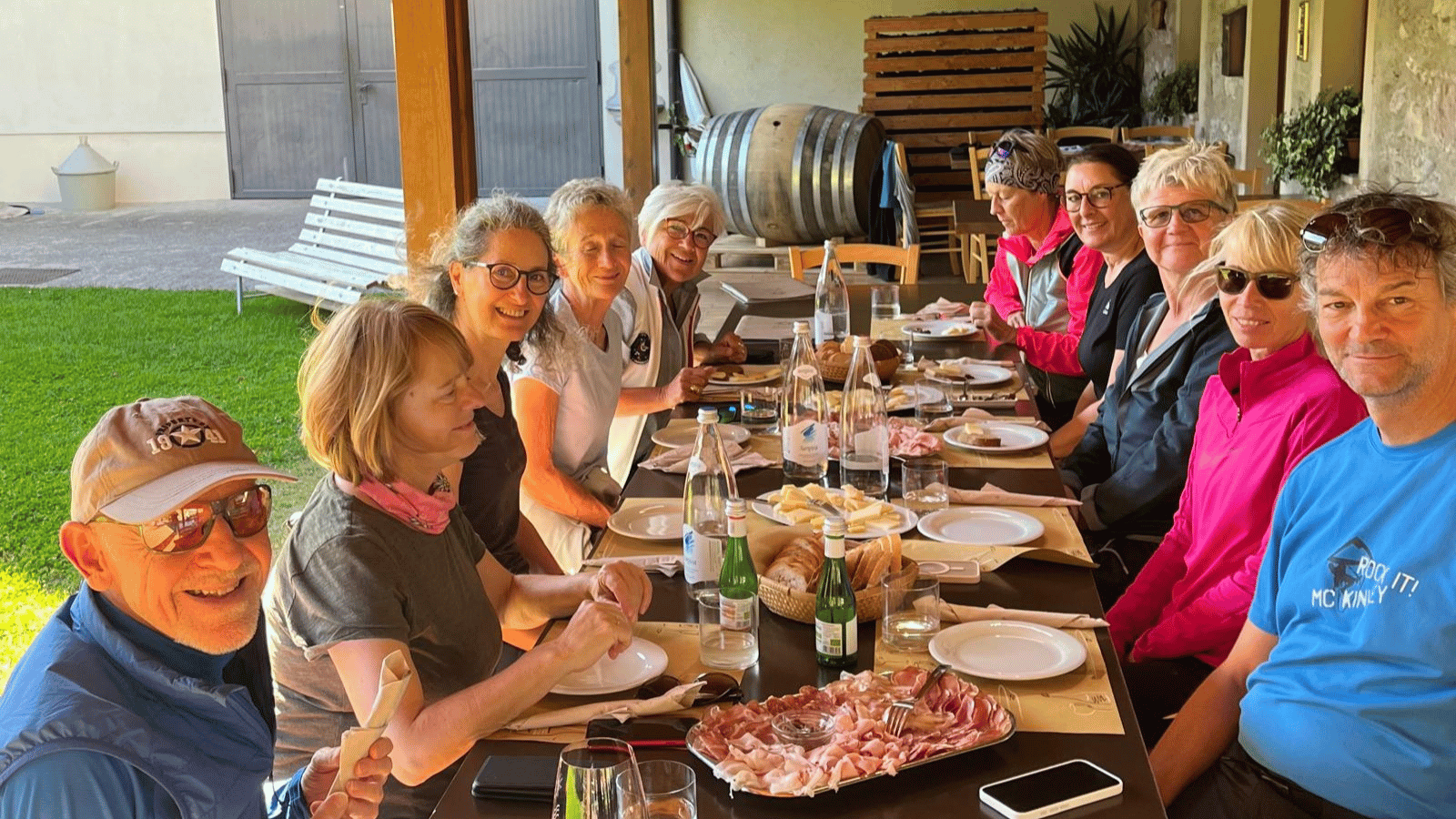 10 Personen sitzen an einem langen Tisch. Vor ihnen Teller und in der Mitte des Tischs Platten mit Wurst/Schinken/Käse sowie Brot. Alle blicken fröhlich in die Kamera.