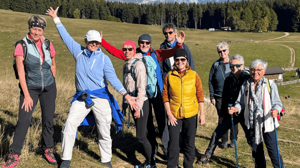 Gruppe von 9 Personen steht im leichten Anstieg vor Feldern und Hintergrund Wald. Zwei Personen, Frau und Mann, recken Arme in die Höhe. Die anderen blicken freundlich in die Kamera. Alle tragen Sportkleidung
